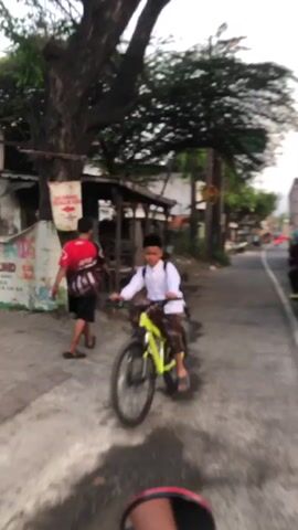 A person wearing a red backpack walks past a small roadside stall. The stall has a wooden frame and is open on one side. A large tree is to the left of the stall, and a paved road is to the right. There are puddles of water on the ground. The video is shaky and appears to be filmed from a moving vehicle.