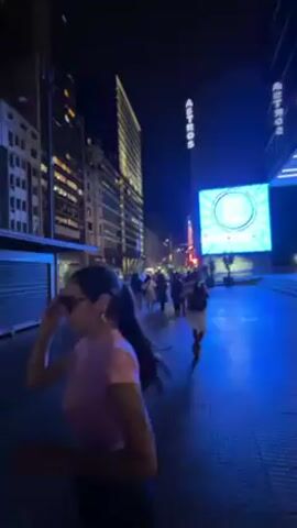 The video shows a night scene in what appears to be a city center. The camera follows a person wearing a red sleeveless shirt and a silver chain around their neck walking with a woman in a pink shirt and sunglasses. They are holding hands. The area is illuminated by streetlights and neon signs, giving it a bright, artificial look. Buildings of varying heights surround the walking area, and other people can be seen strolling around in the distance. Cars occasionally pass on the street. The sidewalk is paved and has markings. Overall, the scene captures a typical urban environment at night.