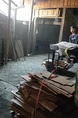 A man is shown weighing stacks of cardboard boxes in a dimly lit workshop. He is wearing a black t-shirt and shorts, and he is using a manual scale. There are several stacks of cardboard boxes around him, as well as some wooden pallets and other materials. The workshop appears to be made of wood and brick, with a tiled floor. Another person is visible in the background, also handling cardboard. The lighting is dim and appears to be natural light coming from an opening in the wall.