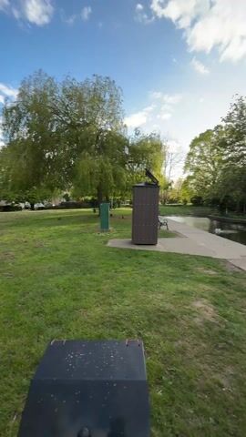 The video captures a serene park scene with a pond under a partly cloudy sky. The camera pans slowly from left to right, revealing various elements of the park.

Initially, the view shows a grassy area on the left with large, green trees. A brown, shed-like structure with a solar panel on its roof stands on the grass, next to a concrete path that leads towards a body of water. A green park bench is visible near the path and the water's edge.

As the camera pans right, the pond becomes the central focus. Several ducks are seen swimming in the calm water, and one duck stands on the concrete path near the pond. A small island of reeds and greenery is situated in the middle of the pond. The pond reflects the surrounding trees and the sky, which features both bright patches and scattered clouds. In the foreground on the right, dark grey metal railings are visible. Lush green trees line the far bank of the pond, and the sun peeks through the clouds, casting a soft light over the scene. The overall impression is one of peaceful natural beauty in an urban or suburban park setting.