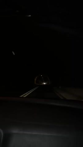 The video shows a view from inside a car driving through a dark tunnel. The tunnel has lights spaced along its ceiling, and a bright light emanates from the end of the tunnel, indicating the exit. The road ahead has white lines marking the lanes.