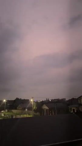 A nighttime video shows a dark, cloudy sky over a suburban landscape. Several houses with illuminated windows are visible, along with streetlights casting beams of light. Two bright points of light, likely stars or planets, are visible in the upper portion of the sky, as are faint streaks of light that appear to be originating from the streetlights. The foreground is dark, obscuring any details. The video appears to be filmed from an elevated position, possibly a balcony or rooftop.