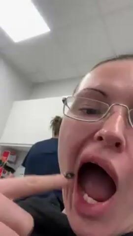 A woman with glasses and black fingernails points inside her mouth in a close-up shot. She opens her mouth wide to show her teeth. Another person wearing a dark shirt is in the background. The setting appears to be an office or utility room. The ceiling has fluorescent lights.