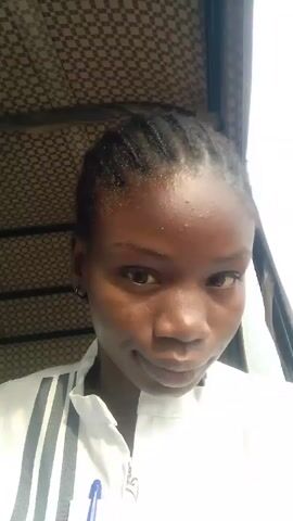 A young woman with braided hair and wearing a white jacket with gray stripes is looking at the camera. The camera is focused on her face and upper body. She appears to be in a vehicle with a checkered pattern ceiling. The background is blurry and light.