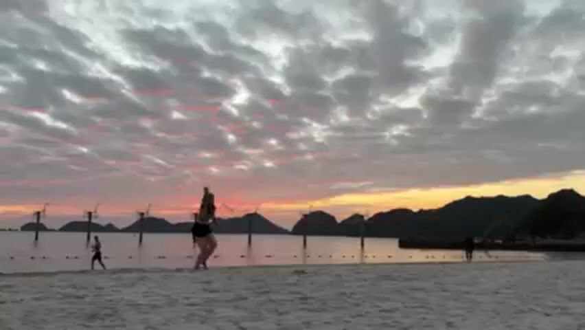 A woman is throwing a frisbee on a beach at sunset. The woman is wearing a black sports bra and black shorts. She throws the frisbee and then picks it up after it falls. There is a pier in the background, and some islands in the distance. The sky is filled with colorful clouds. There are other people on the beach.