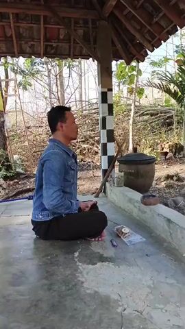 The video shows a man sitting cross-legged on a concrete floor under a thatched roof. He is wearing a blue denim jacket and dark pants. His eyes are closed, and he appears to be meditating. To his right, there is a black clay pot, a small wooden bowl, and a packet of what looks like herbs or spices on the floor. Behind him, a black and white checkered pillar supports the roof. The environment appears to be outdoors, with trees and dry vegetation visible in the background. The lighting suggests it is daytime, possibly morning or late afternoon.