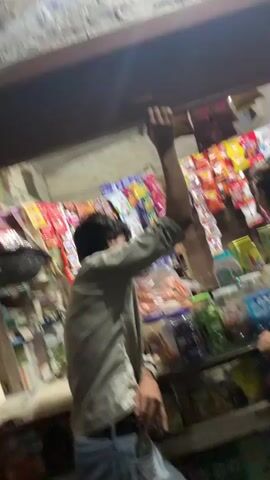 The video shows two young males at a small convenience store. The first male, wearing a light-colored shirt, reaches up towards a shelf above the counter. The second male, in a light green t-shirt, stands behind the counter and gestures towards the shelves stocked with various snacks and products. The environment appears to be an indoor shop with shelves filled with colorful packaged goods. The lighting is dim, suggesting it is evening or indoors with limited light.
