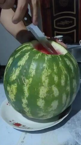 A person's hands are shown cutting a large, striped green watermelon. The watermelon is sitting on a white plate with red floral accents. The person uses a knife to slice a segment from the top of the watermelon, revealing the red flesh and black seeds inside. The top portion of the watermelon is then lifted, showing the cut surface of both the removed piece and the remaining fruit. The person's arm is visible, holding the knife and the watermelon segment. The background is blurred but appears to be an indoor setting with a wall and a door visible. The lighting suggests it is daytime or a well-lit interior.