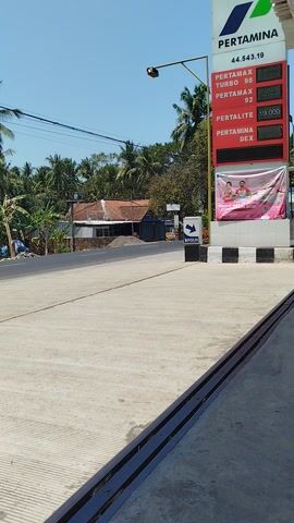This media file depicts an outdoor scene at a Pertamina gas station in Bocor, Indonesia. The time of day is clearly daytime, indicated by the bright sunlight and clear blue sky, suggesting fair weather. The setting is a typical roadside gas station with a concrete forecourt and a visible drainage channel.

The prominent object is the Pertamina price board, displaying fuel types and their corresponding prices. The visible fuel types are Pertamax Turbo 98, Pertamax 92, Pertalite, and Pertamina Dex. The price for Pertamax 92 is shown as 13,800, and Pertamina Dex as 10,000. A banner hangs on the side of the price board, featuring images of two children and text, though the text is partially obscured. There is also a "MASUK" (Entrance) sign pointing to the right.

In the background, a rural Indonesian landscape is visible with houses, lush green trees, and coconut palms, characteristic of the region. There are no people actively engaged in any particular activity that is clearly discernible. The overall impression is one of a bright, sunny day at a local gas station in a tropical environment.