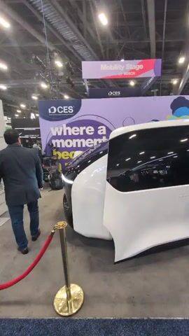 The video shows a white van with its side door open at an indoor event. The van has black accents and the interior is visible. Two men are standing near the van, talking. One man is wearing a black shirt and black pants and the other is wearing a suit. There is a gold stanchion and red rope in front of the van. In the background, there are other people and displays. A sign that says "Mobility Stage" can be seen above the van. The video appears to be taken at a trade show or convention.