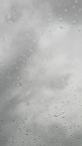 This video shows a close-up view of raindrops on a window. The background is a blurred gray sky, suggesting it is raining. The raindrops are of various sizes, and some are streaking down the glass. The camera is static.