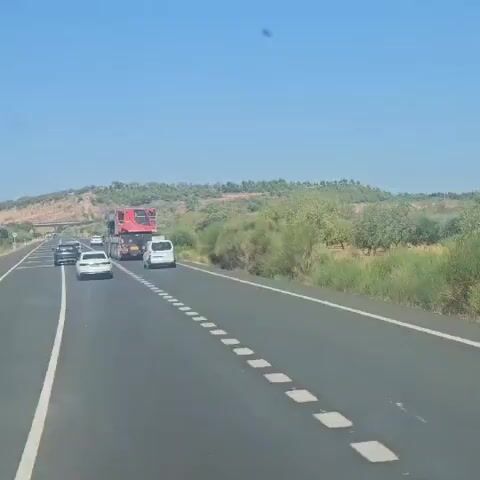 A wide shot captures a highway scene under a clear blue sky. A large red crane is being transported on a flatbed truck, traveling in the right lane. To the right of the truck, a white van is following. Several other cars are visible in the left lane. The landscape is hilly with sparse vegetation and trees. The road has dashed white lines marking the lanes. The video appears to be recorded from a moving vehicle.
