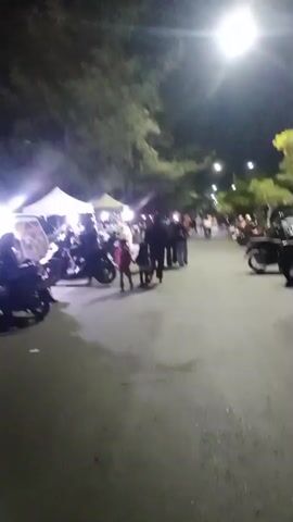 The video shows a nighttime scene on a paved road lined with trees and streetlights. On the left side of the road, there are several tents with bright lights illuminating them, and motorcycles are parked in front of them. A few people are standing near the tents, and some appear to be interacting with each other. Further down the road, on the right side, a large number of motorcycles are parked. People are walking along the road, and some are gathered in groups. The overall atmosphere appears to be that of a night market or a gathering.