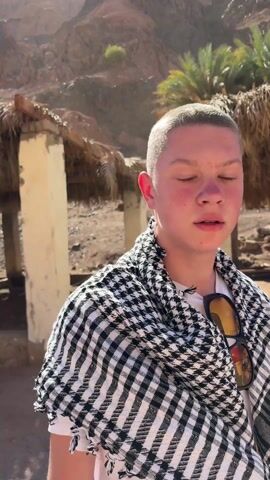 A young person with a buzz cut, wearing a black and white checkered scarf, stands near a straw-roofed structure. The camera pans, revealing a dry, desert landscape with rocky mountains, palm trees, and other tourists. Some tourists wear headscarves, and an old blue jeep is parked nearby. Tourists are gathering near the jeep. The sky is blue with scattered clouds.