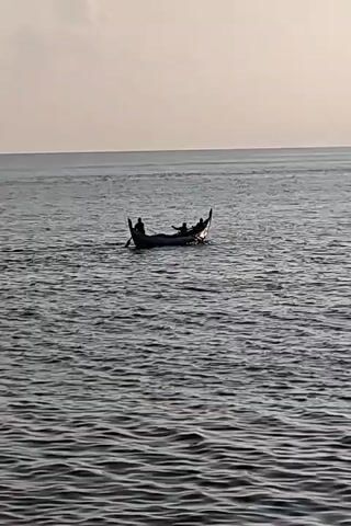 A low-angle shot shows a small, dark-colored boat moving across a body of water. The boat is populated by several silhouetted figures. The water is rippled with small waves. The sky is a pale, even color, suggesting it might be dawn or dusk. The boat appears to be moving away from the viewer. The scene is generally calm.