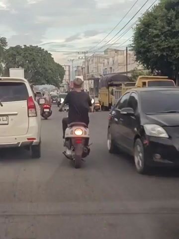 The video shows a street scene with multiple vehicles, including cars and motorcycles, moving in the same direction. The sky is cloudy. On the left side of the road, a white car is visible. In front of it, several motorcycles are moving. Further ahead, a black car is also on the road. On the right side of the road, there are trucks. The camera appears to be mounted on a moving vehicle, likely another car or motorcycle, given the perspective. People are riding the motorcycles, with one person in the foreground wearing a yellow shirt with the number 7. The overall impression is of moderate traffic in an urban or suburban setting.