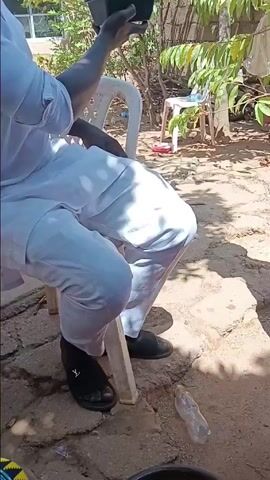 Here is a detailed description of the video:

A man is sitting on a white plastic chair outdoors. He is wearing a light-colored shirt and pants, with black sandals. He holds a black bowl in his left hand. A plastic water bottle lies on the ground near his feet. In the background, there is another white chair, vegetation, and a wall. The ground is a mix of dirt, sand, and some cracked pavement. The scene is partly shaded.