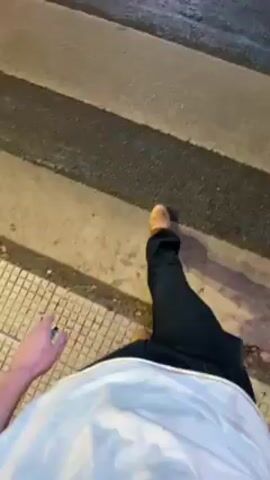 A first-person perspective video shows a person walking across a crosswalk at night. The person is wearing black pants, light brown shoes, and a white shirt. They are holding a small object in their left hand. The crosswalk is made of black and white stripes. The lighting appears to be from streetlights or nearby buildings. The street appears to be wet.