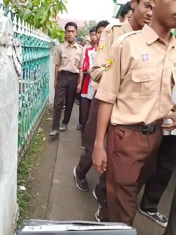 The video shows a group of young people walking along a sidewalk next to a green fence. They are dressed in school uniforms of tan shirts and brown pants or skirts. Some are wearing hijabs. The sidewalk is concrete and there is some vegetation visible alongside the fence. A white building is partially visible in the background. The camera follows the group as they walk.