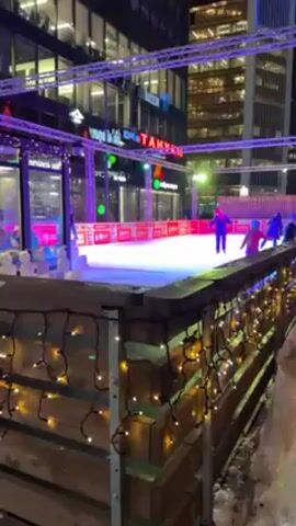The video shows an outdoor ice rink at night, surrounded by tall buildings and decorated with fairy lights. The rink is enclosed by a low wooden fence also adorned with fairy lights. A few people are ice skating on the rink, including children. The rink is illuminated with blue, green, and pink lighting. Across the rink, a sign with Russian text is visible. There is snow around the rink. The camera seems to be stationary.