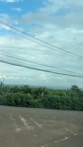 The video pans across a roadside scene with lush green vegetation and a dirt road in the foreground. Overhead, a network of power lines stretches across the frame, supported by utility poles. The sky is partly cloudy with patches of blue. In the distance, more greenery and trees are visible. Towards the end of the video, a white bus with an open door is seen, with a person partially visible inside. The bus has text on its side, including "LUXURY" and what appears to be Vietnamese writing. The overall impression is a rural or suburban landscape.
