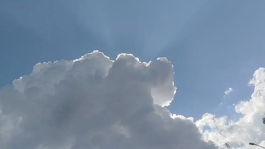 This video is a close-up, upward-facing shot of a large, puffy white cloud against a bright blue sky. Rays of sunlight are visible radiating from behind the cloud. The bottom left and right corners of the frame show the tops of dark power lines and a tall electrical tower. The camera appears to be slightly tilted and the cloud itself is the primary focus of the video.