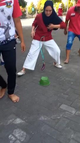 A group of people are playing a game outdoors on a paved area. A woman in the foreground wearing a maroon shirt, white pants, and a black hijab is participating in the game. She has a string tied around her waist and a purple object, possibly an eggplant, is hanging from the string. She is bending over, attempting to get the object into a green plastic bowl placed on the ground in front of her. Another person wearing a red shirt and black pants is standing to her left, and a person in a red shirt and blue jeans is to her right. Other people in red shirts are visible in the background, along with trees and buildings. The ground is paved with interlocking bricks.