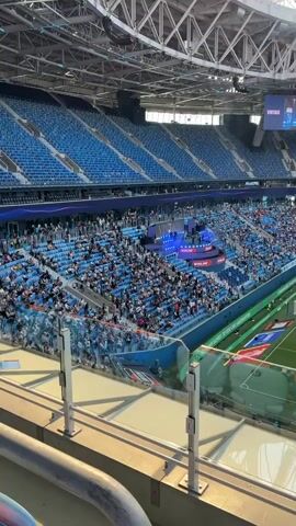 The video pans across a wide view of a modern, outdoor soccer stadium with tiered blue seating. The stands are partially filled with spectators. In the center of the field, several players in light blue uniforms are visible warming up. A stage area with blue lighting and a screen is set up on one side of the field. Above the field, a large screen displays an image or video. The stadium has a visible metal roof structure. The lighting suggests it is daytime, possibly late afternoon or early evening.