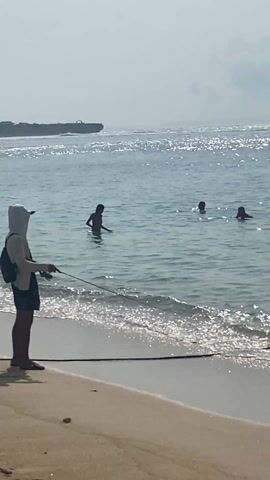 The video shows a sunny beach scene. A person wearing a white hooded shirt, dark shorts, sandals, and a backpack stands on the sandy beach, holding a fishing rod and casting a line into the ocean. There are three people swimming in the water some distance from the shore. Further out at sea, there is a dark, rocky formation visible on the horizon. The water sparkles with sunlight.