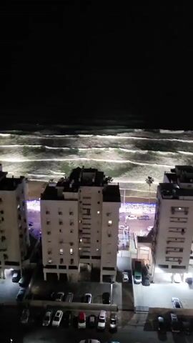 The video shows a nighttime cityscape next to an ocean. Two tall, white apartment buildings stand in the foreground, their windows lit with warm light. A parking lot filled with cars is located at the base of the buildings. In the background, the ocean is visible, with waves illuminated by the city lights, creating bright streaks on the water. A palm tree stands near the beach, surrounded by bright blue lights. The night sky is dark and clear. The camera slowly pans to the right, revealing more of the building and the coastline.