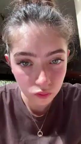 A close-up shot shows a young woman with her hair pulled back, wearing a brown t-shirt and a delicate necklace. She is applying brow gel to her eyebrow with a small wand applicator. Sunlight illuminates her face, highlighting the texture of her skin. The background is out of focus, but hints at an indoor setting with glimpses of a ceiling fan and wooden elements.