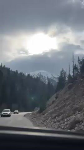 The video shows a scenic mountain landscape. A car drives on a paved road with a snowy mountain visible in the distance under a cloudy sky. The camera pans up to show more of the snow-covered mountain with evergreen trees in the foreground. The mountain appears partially bare, showing rock formations as well as snow. The audio captures the sound of wind.