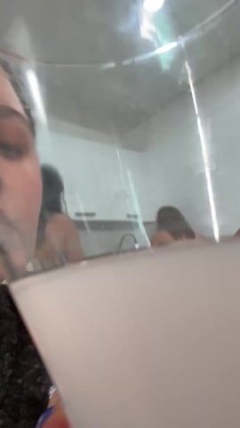 A woman with dark hair pulled back and blue and white striped nails holds a tall glass of white liquid up to her face and takes a sip. Two other women are in the background, in a bright white kitchen with white cabinets and a stainless steel sink. One woman is washing dishes and the other is setting plates.
