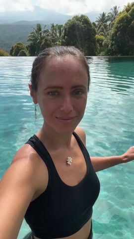A woman is standing in a turquoise-colored swimming pool. She is wearing a black tank top and a necklace with a white pendant. Her hair is pulled back, and she is wearing dangling earrings. She smiles at the camera. The pool appears to be an infinity pool, as the water flows over the edge. Behind the pool, there are green trees and distant hills under a cloudy sky.