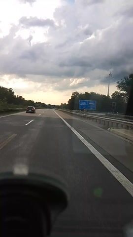 The video shows a view from inside a vehicle traveling on a highway. The camera is mounted on the windshield, and the perspective is angled to the right. The road is asphalt with white lane markings. On the left side of the highway, there is a guardrail, trees, and a cloudy sky. There are signs in the distance. A few cars are visible on the highway ahead. The video is filmed during the day.