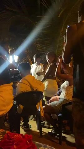The video shows a group of people gathered outside at night. Several people are standing and dancing, while a few others are seated in chairs. One person is filming the gathering with a smartphone. The lighting is provided by an artificial light source, possibly a streetlight, creating bright spots and shadows. Palm fronds are visible in the background, adding to the outdoor atmosphere. Some individuals are wearing light-colored clothing, while others are in darker attire, and one is wearing camouflage pants. There are blankets on the ground and a festive vibe to the gathering.