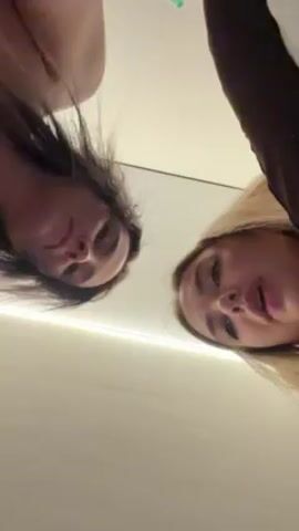 The video shows two women looking down into the camera. The woman on the left has long, dark hair and the woman on the right has blonde hair and is wearing a brown top. The background is a white surface. The women are both smiling and talking, with the blonde woman appearing to mouth words.