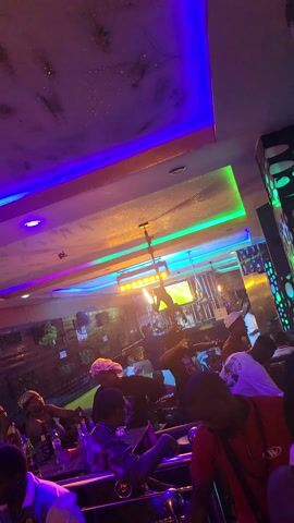 This video captures an indoor scene, likely a lounge or shisha bar, at night in Obosi, Nigeria. The setting is dimly lit with dynamic, colorful LED strip lighting, predominantly green, blue, orange, purple, and red, illuminating the walls and ceiling.

Numerous people, mostly young adults, are visible socializing. They are seated at various tables, some standing, engaged in conversations. Many tables feature shisha pipes with multiple hoses, indicating that shisha smoking is a primary activity. People are seen holding drinks in bottles and glasses.

The decor includes walls with a marbled pattern and sections with metallic, dotted, or geometric designs. Ceiling fans are visible, circulating air. The atmosphere appears lively and social, with background music audible throughout the video. Reflections of the neon lights are visible on surfaces, creating a vibrant ambiance.

No explicit text is clearly legible, though there might be faint signage in the background or text on bottles that is too blurry to decipher. The focus is on the social interaction and the modern, colorfully lit environment of the establishment.