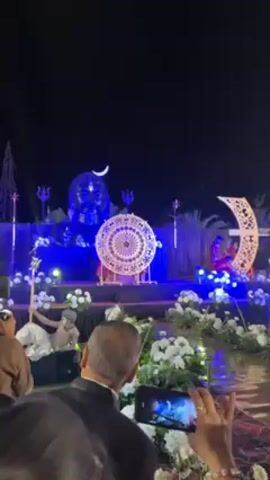 The video shows a nighttime outdoor event with an elaborate stage set. On the stage, there is a large statue of the face of Shiva, an ornamental crescent moon, a decorative circular object in the center, and floral arrangements. A person can be seen standing behind the circular object. The scene is illuminated with blue stage lighting. In front of the stage, a crowd of people is watching and recording the event with their smartphones. The environment appears to be an open-air venue prepared for a special occasion.