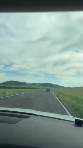 The video captures a view from inside a moving car, looking out through the windshield onto a two-lane asphalt road. The road stretches towards a distant horizon, lined with green grassy fields and rolling hills. A single dark-colored car is visible ahead, also traveling on the same road. The sky is mostly filled with a dynamic cloud cover, with patches of blue sky peeking through, suggesting a daytime scene with varied weather. The overall impression is that of a road trip through a rural, possibly steppe or grassland, landscape. The car interior is partially visible in the foreground, including the dashboard. There are no people visible outside the car, and no text is present in the video. The location context provided is Znamenka, Russia, which suggests the setting is a rural area in that region.