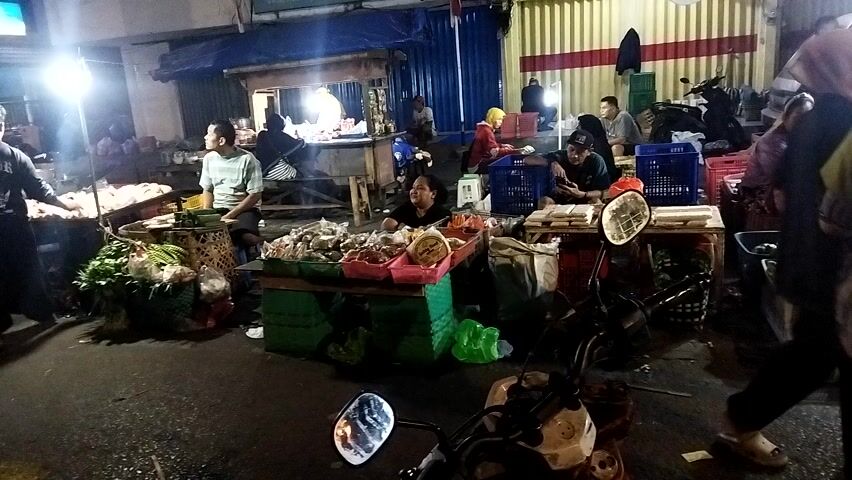 The video shows an outdoor market at night. There are several stalls selling various items, including fruits, vegetables, and possibly seafood. People are visible at the stalls, some interacting with vendors, others appearing to be customers. A motorcycle is parked in the foreground. The market appears to be active, with lights illuminating the area.