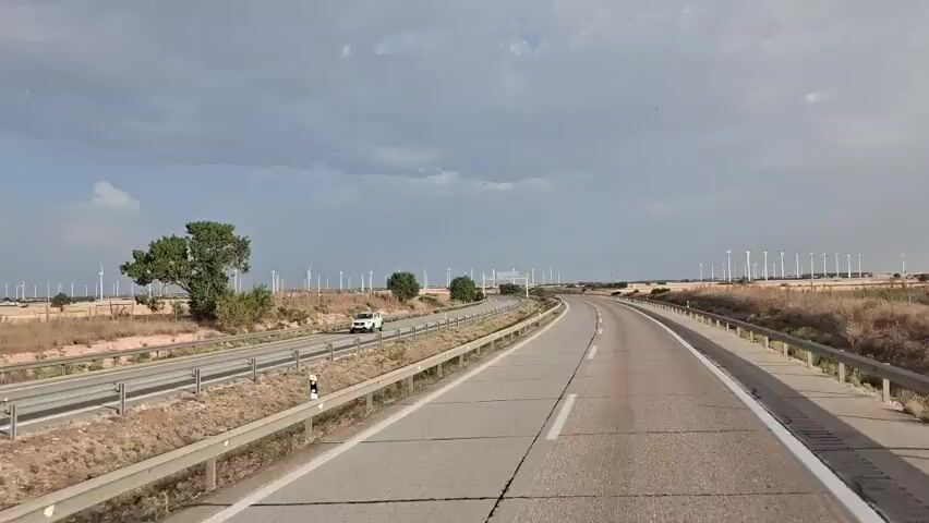 The video shows a view from a vehicle traveling on a highway with a dual carriageway. On the left, there is a parallel road and a grassy median. On the right, there is a field of dry grass and scattered trees. In the distance, a large wind farm with numerous wind turbines is visible across the landscape under a cloudy sky. A white SUV is seen on the left-hand road, moving in the opposite direction. The highway has clear lane markings, and guardrails are present along the sides. The scene appears to be in a rural or semi-arid environment.