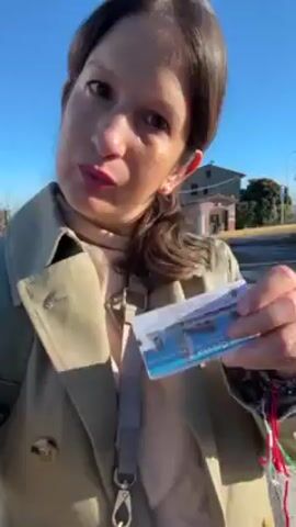 A woman in a tan trench coat and a light-colored turtleneck stands outside during the day. The sky is clear and blue. She is holding a small plastic card with a white background and blue and gray designs. She purses her lips and looks directly at the camera. She wears a strap across her chest, a wedding ring, and has her hair pulled back. There is a road in the background. A backpack strap is visible on her shoulder. A sound resembling church bells is audible.