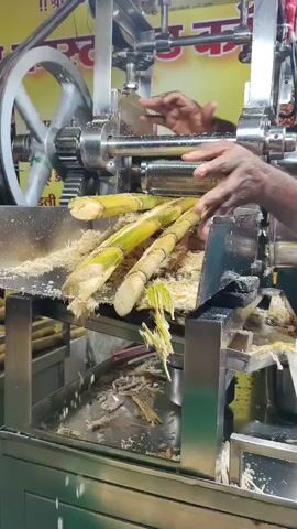 A person is operating a sugarcane juicing machine. The machine is made of stainless steel and has large rollers. The person is feeding sugarcane stalks into the machine, and the juice is being extracted and collected in a tray. The sugarcane stalks are green and appear to be fresh. The environment seems to be an outdoor market or a roadside stall. There is text on a banner behind the machine, which appears to be in Hindi.