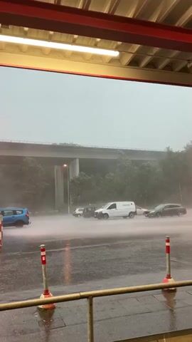 The video captures a severe rain and possibly hail storm during the daytime in Neunkirchen, Germany. The scene is viewed from under a covered area, possibly a building or an underpass, as evidenced by the red overhead structure and fluorescent light. In the foreground, two red-and-white striped safety posts are visible.

A street runs across the mid-ground, where several vehicles are navigating through exceptionally heavy rainfall. Large amounts of water are splashing high from the road surface due to the intensity of the rain and the movement of cars. A blue SUV is seen on the left side of the road, and a white van, with "Hermes" visible on its side, is in the center. Other dark-colored cars are also present further down the street.

In the background, a large concrete highway or bridge overpass stretches across the frame, with trees and dense foliage visible beneath it. A street lamp with an orange glow adds a point of light to the dim, overcast conditions.

The audio features the distinct, overwhelming sound of heavy rain and strong wind. A very loud, sharp cracking sound, characteristic of thunder or a close lightning strike, occurs early in the video, further emphasizing the extreme weather conditions. The entire scene conveys a sense of intense storm and challenging driving conditions.