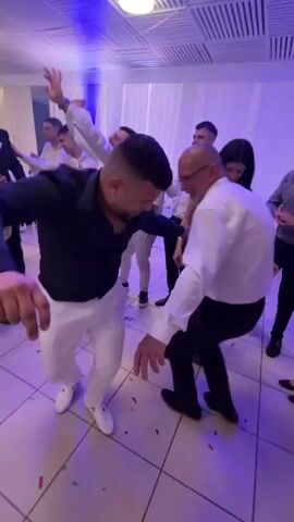 People are dancing at a wedding reception. Men in white suits and shirts, and a man in a navy shirt and white pants dance in a circle. Some wave white napkins. A saxophonist in a black suit and a drummer are nearby. A woman in a black dress and another in a white dress stand near the dancers. The room has white curtains, a light-colored tile floor with confetti, and a modern ceiling.