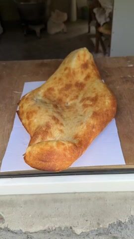 A large, golden-brown flatbread sits on a white square paper on a wooden counter. A man's hand reaches into the frame from the top right, then retreats. A woman's hand wearing a ring briefly touches the paper beneath the bread. The background includes a glimpse of what looks like a shop interior with a large metal bowl or pot and a chair visible. The counter is set in front of what appears to be a window frame or ledge. The video is taken at a medium-close range and is well-lit.