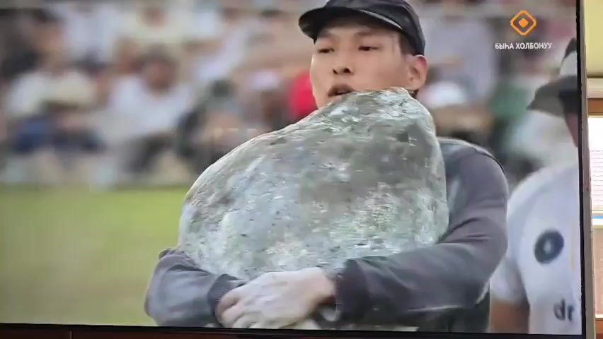 A man wearing a grey long-sleeved shirt, dark jeans, and a black baseball cap is carrying a large, rough, gray rock. His arms are wrapped around the rock, and he is holding it close to his chest. He appears to be exerting himself, with his mouth slightly open. He is walking on a grassy field. Another man, wearing a white t-shirt and a hat, is walking behind him and to the right, holding two colorful poles. In the background, there is a large crowd of people watching, and some tents or structures. A triangular marker is visible in the field to the left of the man carrying the rock. The video appears to be a recording of an outdoor event or competition.