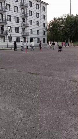 A group of people are gathered in an open, paved area in front of a white apartment building. Several people, including men and women, are spread out on the ground, some are standing and watching. Two people appear to be playing a game, possibly a sport, as they are standing on opposite sides of a line marked by two red poles. A stroller is parked to the right of the group. The sky is overcast and the lighting is dim. The environment appears to be a residential area, possibly a playground or courtyard.