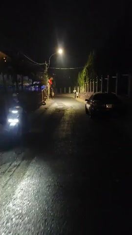 The video depicts a dimly lit street at night. A single streetlamp illuminates the center of the frame, casting light down the road. The asphalt is cracked and patched, suggesting age and wear. On the left side of the street, a wooden fence and a building adorned with colorful lights can be seen. On the right, a dark car is parked beside a stone wall lined with evergreen trees. In the distance, several people are walking. The scene has a quiet, residential feel.